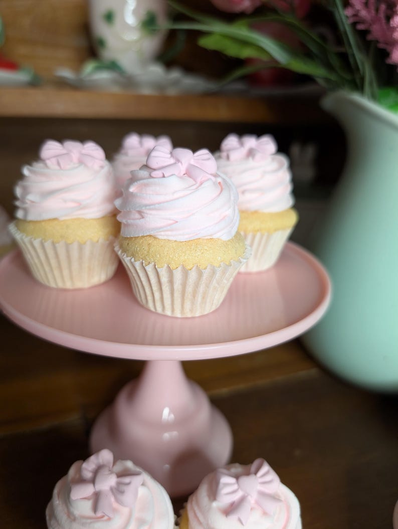 May include: Pink frosted cupcakes with bow decorations on a pink cake stand. The cupcakes have yellow cake bases and are in white paper liners. A light green vase with flowers is in the background.