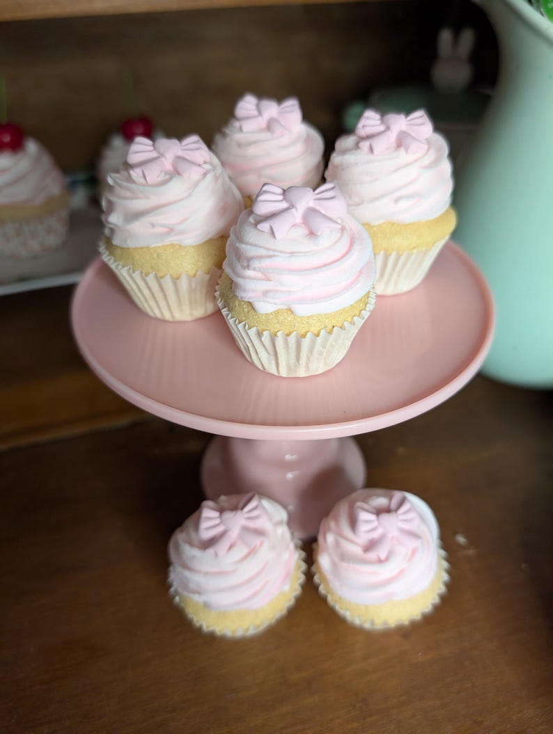 May include: A collection of cupcakes with pink frosting and bow-shaped decorations. The cupcakes are displayed on a pink cake stand. The cupcakes have yellow cake bases and are in white paper liners. Some cupcakes are in the background.