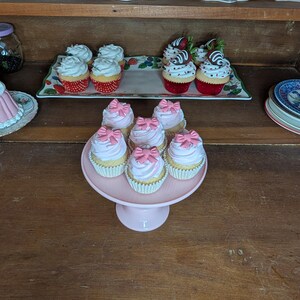 May include: A display of cupcakes with pink frosting and bow decorations on a pink cake stand. Other cupcakes with white frosting and strawberry toppings are on a decorative tray. Teacups and saucers are on a shelf above.
