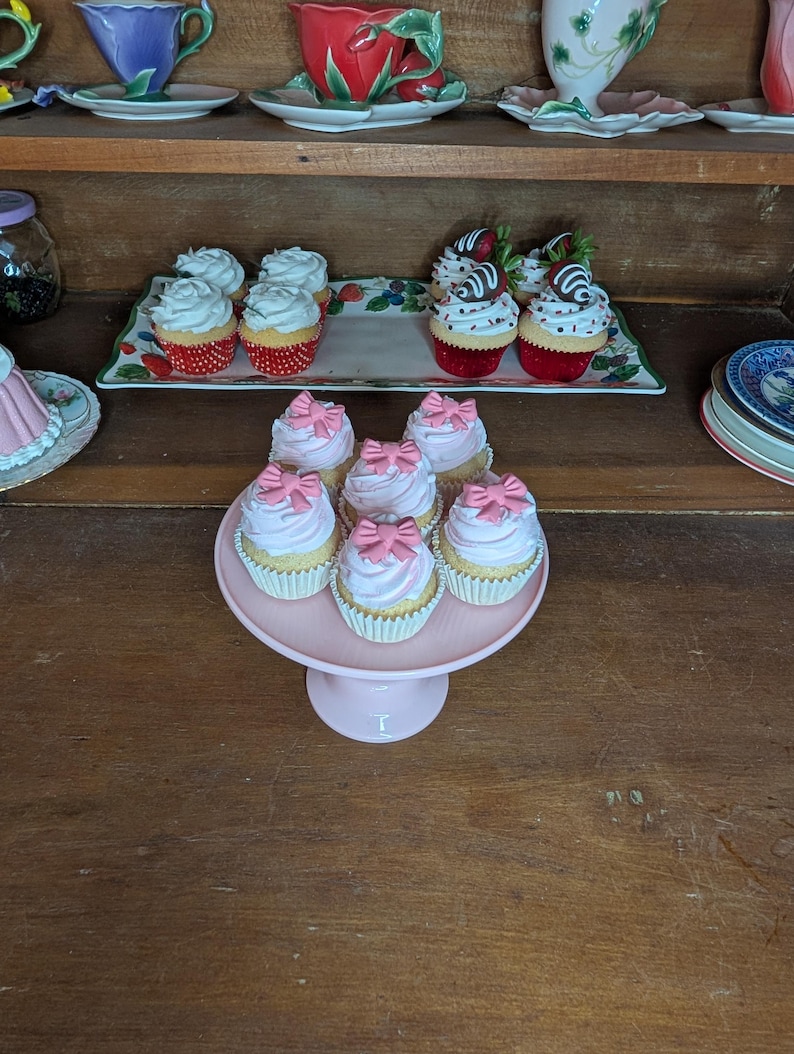 May include: A display of cupcakes with pink frosting and bow decorations on a pink cake stand. Other cupcakes with white frosting and strawberry toppings are on a decorative tray. Teacups and saucers are on a shelf above.