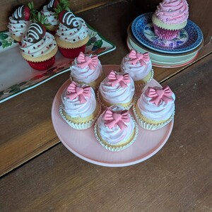 May include: A collection of decorative cupcakes with various toppings. Some cupcakes have pink frosting and pink bow decorations, while others feature chocolate-covered strawberries. The cupcakes are arranged on plates and trays.