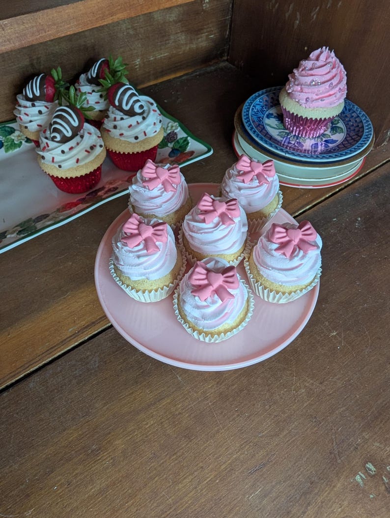 May include: A collection of decorative cupcakes with various toppings. Some cupcakes have pink frosting and pink bow decorations, while others feature chocolate-covered strawberries. The cupcakes are arranged on plates and trays.