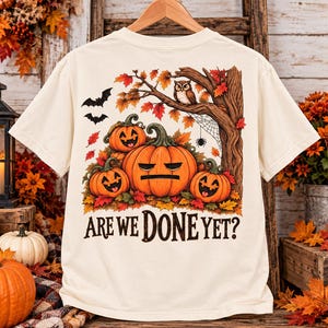 May include: A cream-colored t-shirt featuring a graphic print of four orange pumpkins with carved faces, an owl perched on a tree branch, bats, and a spider web. The text "ARE WE DONE YET?" is printed in black below the pumpkin illustration.