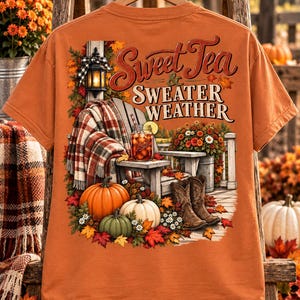 May include: An orange t-shirt featuring a graphic print of a wooden chair with a plaid blanket, a glass of iced tea, pumpkins, cowboy boots, and autumn leaves. The text reads Sweet Tea & SWEATER WEATHER in a decorative font style.