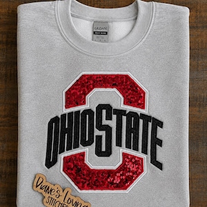 May include: A light gray crewneck sweatshirt featuring a large embroidered Ohio State logo with red sequin accents and black lettering. The garment includes a Gildan brand label at the inner neckline and a wooden sign reading Diane's Loving Stitches.