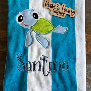 May include: A blue and white striped beach towel with an embroidered cartoon turtle and the name "Santino" in navy blue. A wooden tag with the text "Diane's Loving Stitches" is attached.