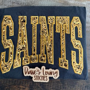 May include: A black t-shirt featuring the word SAINTS in large, gold sequined block letters with a gold stitched border. A small, light-colored wooden sign with the text Diane's Loving STITCHES is placed over the bottom of the shirt.