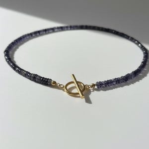 Op de afbeelding: Een delicate ketting met kleine, donkerblauwe kralen. De ketting is voorzien van een goudkleurige toggle sluiting. De kralen lijken gelijkmatig van grootte en aan elkaar geregen, wat een eenvoudig maar elegant ontwerp creëert.