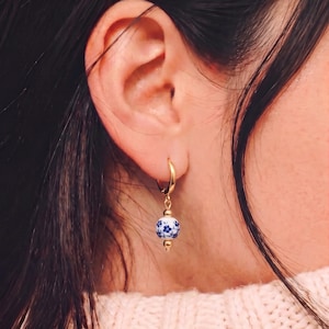 Op de afbeelding: Een gouden oorring met een hangende, bolvormige kraal met een wit en blauw bloemmotief. De kraal is aan de onderkant voorzien van een klein goudkleurig metalen accent en hangt aan een gepolijste gouden ring die door de oorlel is bevestigd.
