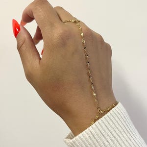 May include: A gold-toned hand chain bracelet featuring a delicate, flat-link chain design. The jewellery piece connects a ring component worn on the finger to a bracelet component that encircles the wrist, creating a continuous decorative link.