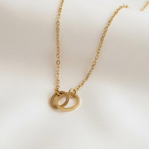 May include: A gold necklace featuring two interlinked rings as a pendant. The chain and rings have a simple, elegant design. The necklace is displayed against a white background.