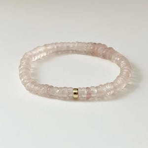 Puede incluir: Una delicada pulsera con cuentas de piedras preciosas facetadas de color rosa pálido. La pulsera está ensartada con una cuenta espaciadora dorada. Las cuentas tienen una calidad translúcida y la pulsera está sobre un fondo blanco.