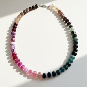 May include: A multi-coloured beaded necklace with a gradient of colours, from dark brown to pink and teal. The necklace is made of small, round beads and has a silver clasp. The beads are approximately 0.6 cm in diameter.