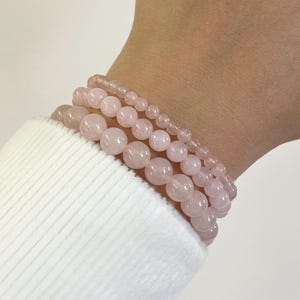 Könnte beinhalten: Vier Armbänder aus Rosenquarz-Perlen in verschiedenen Größen sind auf einem Handgelenk gestapelt. Die Armbänder sind zartrosa und scheinen aus polierten Steinen zu bestehen. Das Handgelenk ist von einem weißen Langarmshirt bedeckt.