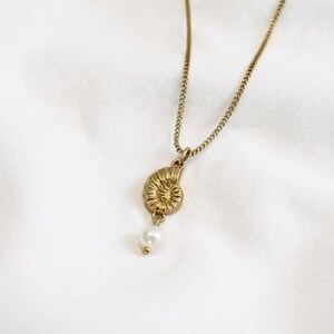 May include: Gold-toned necklace featuring a detailed ammonite pendant with a small, dangling pearl. The chain has a delicate curb link design. The pendant and chain are set against a white background.