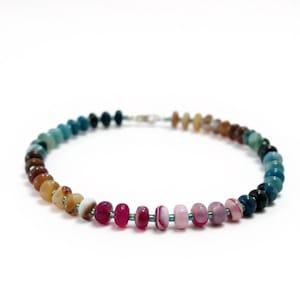 May include: A colourful beaded bracelet displaying a gradient of hues, including blues, greens, browns, yellows, pinks, and purples. The beads are strung on a thin, teal cord and fastened with a silver clasp. A stylish piece of jewellery.