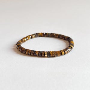 Peut inclure: Un bracelet circulaire composé de petites perles facettées en forme de disque en œil-de-tigre dans des tons marron et doré. Le bracelet comporte une perle unique en métal doré, lisse et polie, intégrée dans la séquence de perles en pierre.