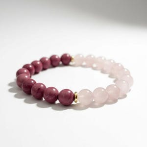 May include: A beaded bracelet featuring two types of gemstones. The left side has dark pink beads, while the right side has light pink beads. A small gold-colored spacer separates the two bead types. The bracelet is on a white surface.