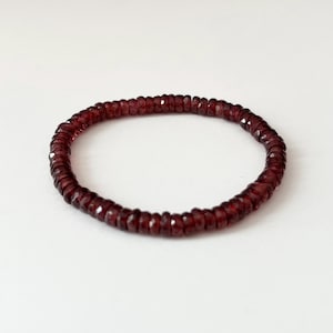 May include: A deep red beaded bracelet. The bracelet is made of small, faceted beads strung together to form a circular shape. The beads have a rich, dark red color, and the bracelet is set against a white background.