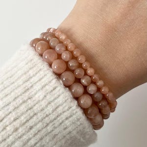 Könnte beinhalten: Vier Armbänder aus pfirsichfarbenen, runden Edelsteinperlen sind auf einem Handgelenk gestapelt. Die Perlen variieren in der Größe und erzeugen einen geschichteten Effekt. Das Handgelenk ist teilweise von einem cremefarbenen Pullover bedeckt.