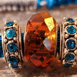 May include: A close-up of a turquoise braided bracelet with a large, faceted amber-colored crystal bead in the center. Silver-toned beads with turquoise-colored stones flank the central bead. The bracelet has a detailed, woven design.
