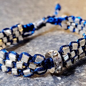 May include: A woven bracelet featuring silver-colored square beads and dark blue cord. The bracelet has a sliding knot closure and a decorative silver ring with clear crystals. The bracelet is approximately 7 inches in length.