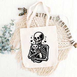 May include: A white tote bag with a black illustration of a skeleton holding a cat. The skeleton has a skull with a toothy grin and the cat has a surprised expression. There are small hearts and stars around the skeleton and cat.