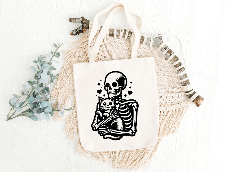 May include: A white tote bag with a black illustration of a skeleton holding a cat. The skeleton has a skull with a toothy grin and the cat has a surprised expression. There are small hearts and stars around the skeleton and cat.