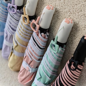 Puede incluir: Una colección de paraguas compactos de colores pastel. Los paraguas cerrados presentan diseños a rayas y asas a juego. Los colores incluyen rosa, amarillo y azul. Los extremos blancos están adornados con huellas de patas. Cada paraguas tiene un diseño único.