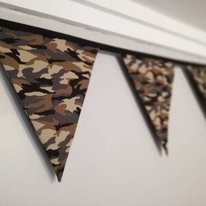 Puede incluir: Una guirnalda de tela con estampado de camuflaje con banderas triangulares. La guirnalda est&aacute; colgada del techo.