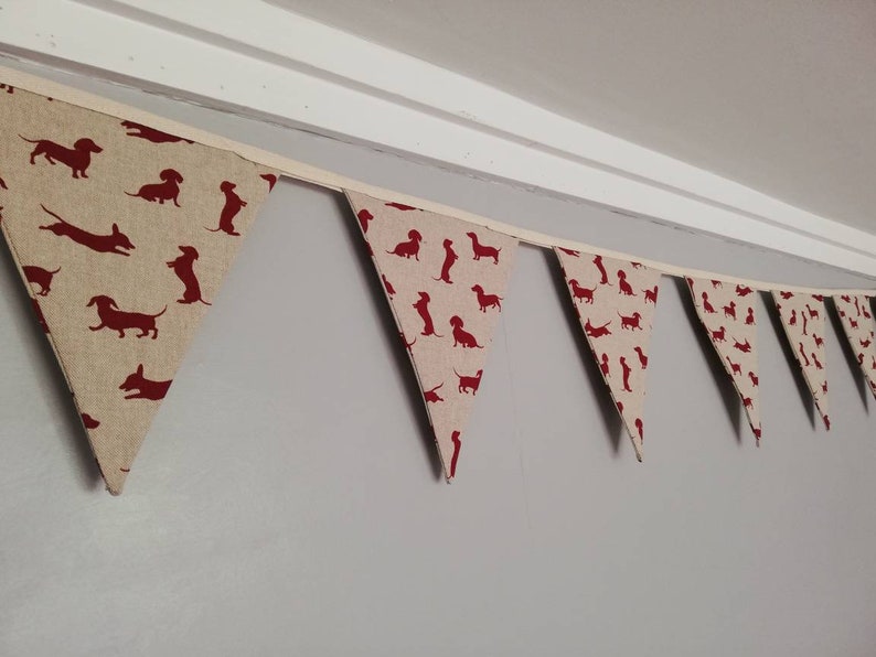May include: A bunting with a string of triangular flags featuring a repeating pattern of red dachshund silhouettes on a beige background.