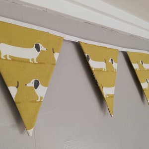 May include: A yellow and white bunting featuring a dachshund design. The bunting is made of fabric and has a white ribbon running through it.
