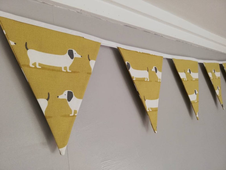 May include: A yellow and white bunting featuring a dachshund design. The bunting is made of fabric and has a white ribbon running through it.