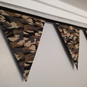 Puede incluir: Una guirnalda de banderas de tela con estampado de camuflaje, colgando de un techo blanco. Las banderas son triangulares y tienen un borde negro.