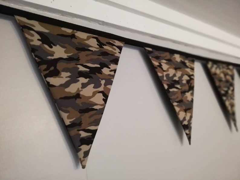 Puede incluir: Una guirnalda de banderas de tela con estampado de camuflaje, colgando de un techo blanco. Las banderas son triangulares y tienen un borde negro.
