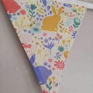 May include: A white fabric bunting with a colorful floral pattern and bunny silhouettes. The bunting is made of cotton and has a white trim. The bunnies are in shades of pink, yellow, and purple.