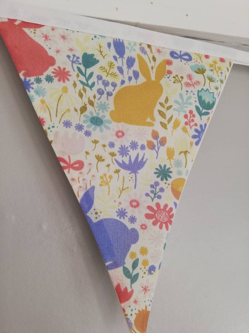 May include: A white fabric bunting with a colorful floral pattern and bunny silhouettes. The bunting is made of cotton and has a white trim. The bunnies are in shades of pink, yellow, and purple.
