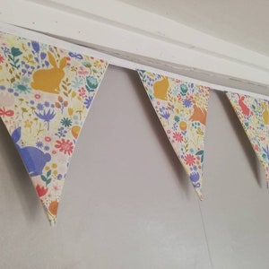 May include: A bunting with a white background and a colorful floral pattern featuring bunnies. The bunting is made of fabric and has a white trim.