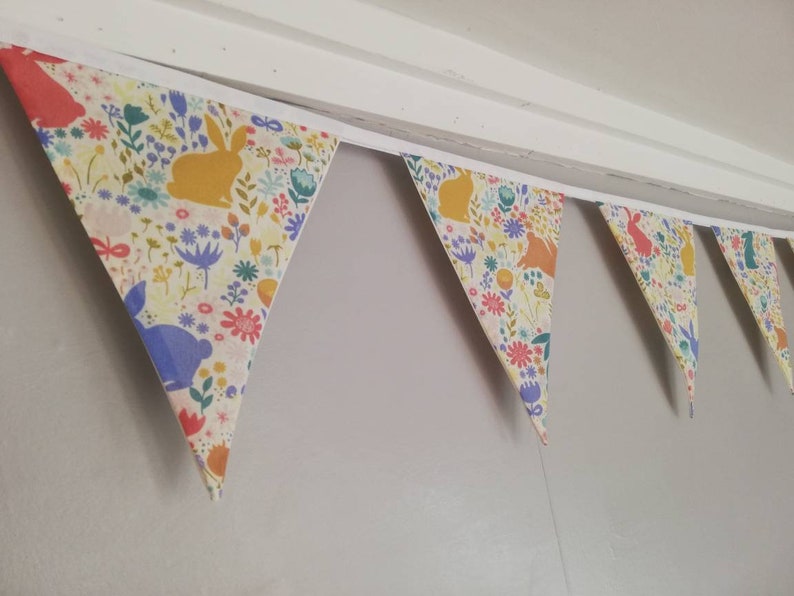 May include: A bunting with a white background and a colorful floral pattern featuring bunnies. The bunting is made of fabric and has a white trim.