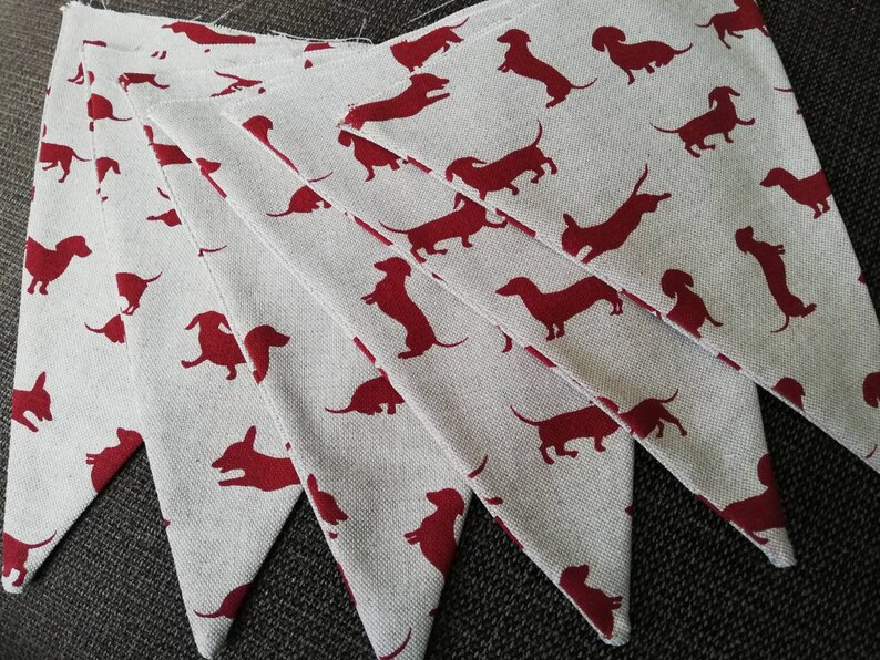 May include: A set of seven white fabric pennants with red dachshund silhouettes. The pennants are sewn together to create a garland.