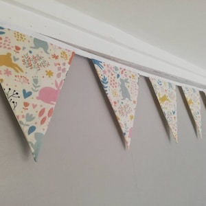 May include: A bunting with a white background and a colorful pattern of bunnies, flowers, and hearts. The bunting is made of fabric and is strung together with white string.