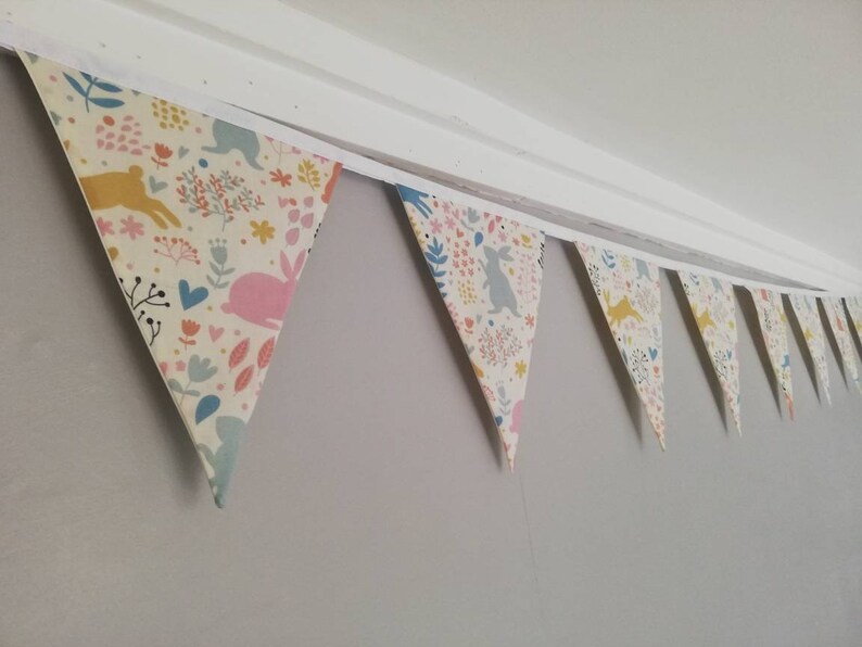 May include: A bunting with a white background and a colorful pattern of bunnies, flowers, and hearts. The bunting is made of fabric and is strung together with white string.