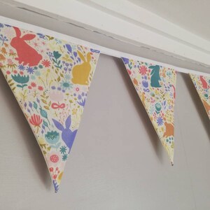 May include: A colorful fabric banner with a repeating pattern of bunnies and flowers. The banner is made of a light-colored fabric with a white trim. The banner is hung on a wall.