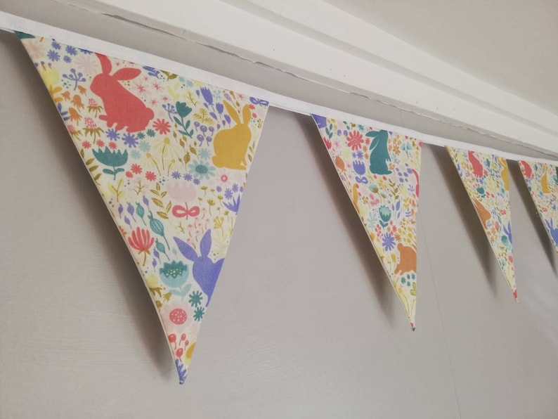 May include: A colorful fabric banner with a repeating pattern of bunnies and flowers. The banner is made of a light-colored fabric with a white trim. The banner is hung on a wall.
