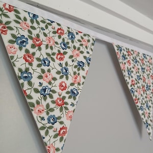 May include: Fabric bunting with a floral pattern in red, blue, and pink on a white background. The triangular flags are strung together, creating a decorative garland against a white and grey background.