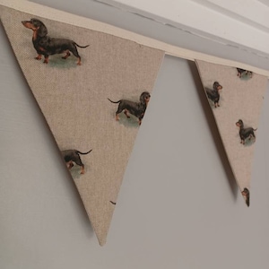 May include: A beige fabric bunting with a repeating pattern of black dachshund dogs. The bunting is strung up on a white wall.