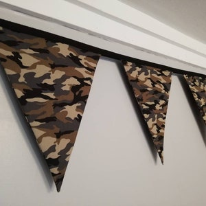 Puede incluir: Una guirnalda de tela con estampado de camuflaje con banderas triangulares. La guirnalda est&aacute; enhebrada en una cinta negra y cuelga de un techo blanco.
