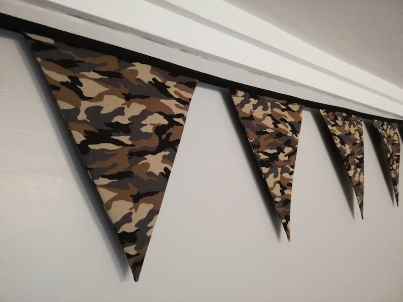 Puede incluir: Una guirnalda de tela con estampado de camuflaje con banderas triangulares. La guirnalda est&aacute; enhebrada en una cinta negra y cuelga de un techo blanco.