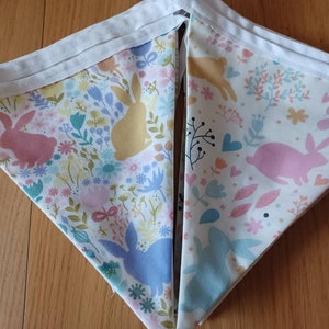 May include: A white fabric bunting with a repeating pattern of pink, yellow, and blue bunnies and floral designs. The bunting is made of cotton and has a white trim.