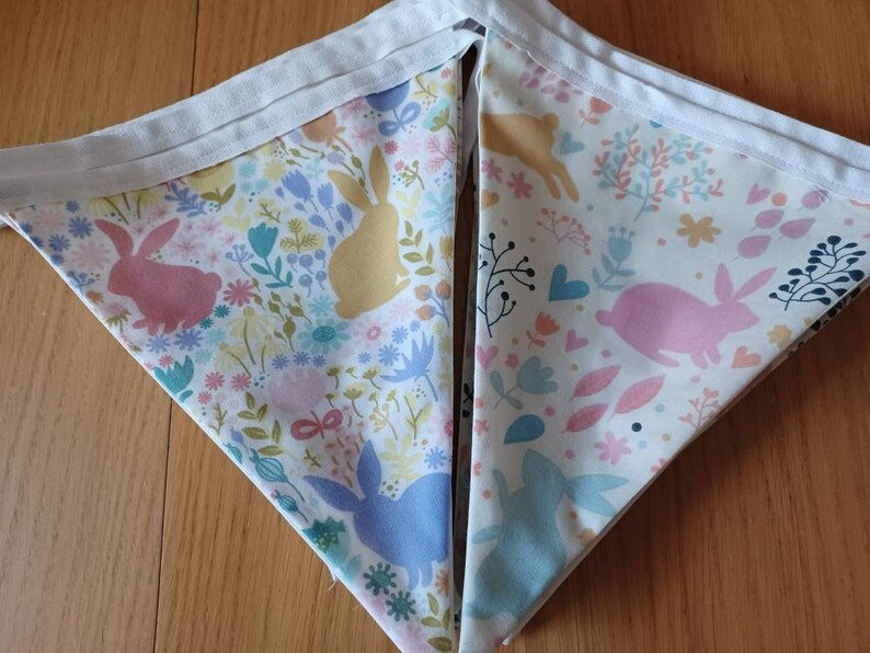 May include: A white fabric bunting with a repeating pattern of pink, yellow, and blue bunnies and floral designs. The bunting is made of cotton and has a white trim.